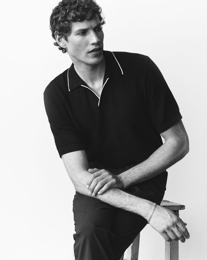Black and white portrait of Paul Hupfer seated on a wooden stool wearing a dark knit polo shirt against a white backdrop.