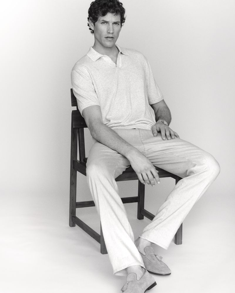 Monochrome studio portrait of a male model seated on a dark wooden chair wearing a light polo shirt and trousers against a pale background