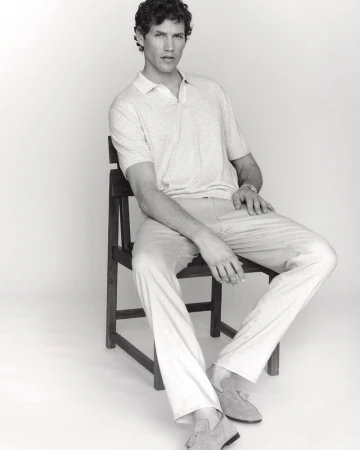 Monochrome studio portrait of a male model seated on a dark wooden chair wearing a light polo shirt and trousers against a pale background