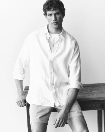 A man in a white button-down shirt and shorts leaning against a wooden table in a studio setting.
