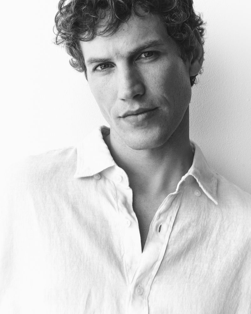 Black and white portrait of a man with short curly hair wearing an unbuttoned white linen shirt against a plain light background.