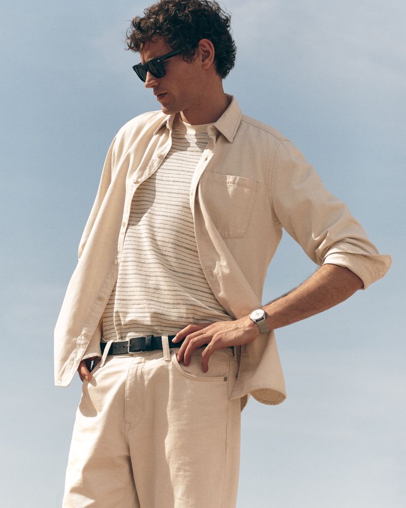 Model Paul Hupfer wearing a light beige overshirt, striped tee, and beige trousers with sunglasses against a pale sky.
