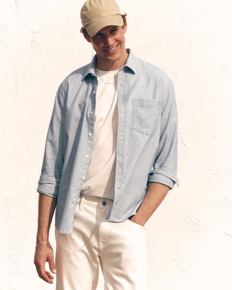 Model Paul Hupfer wears a light blue button-down shirt, white t-shirt, and beige cap against a bright textured wall.