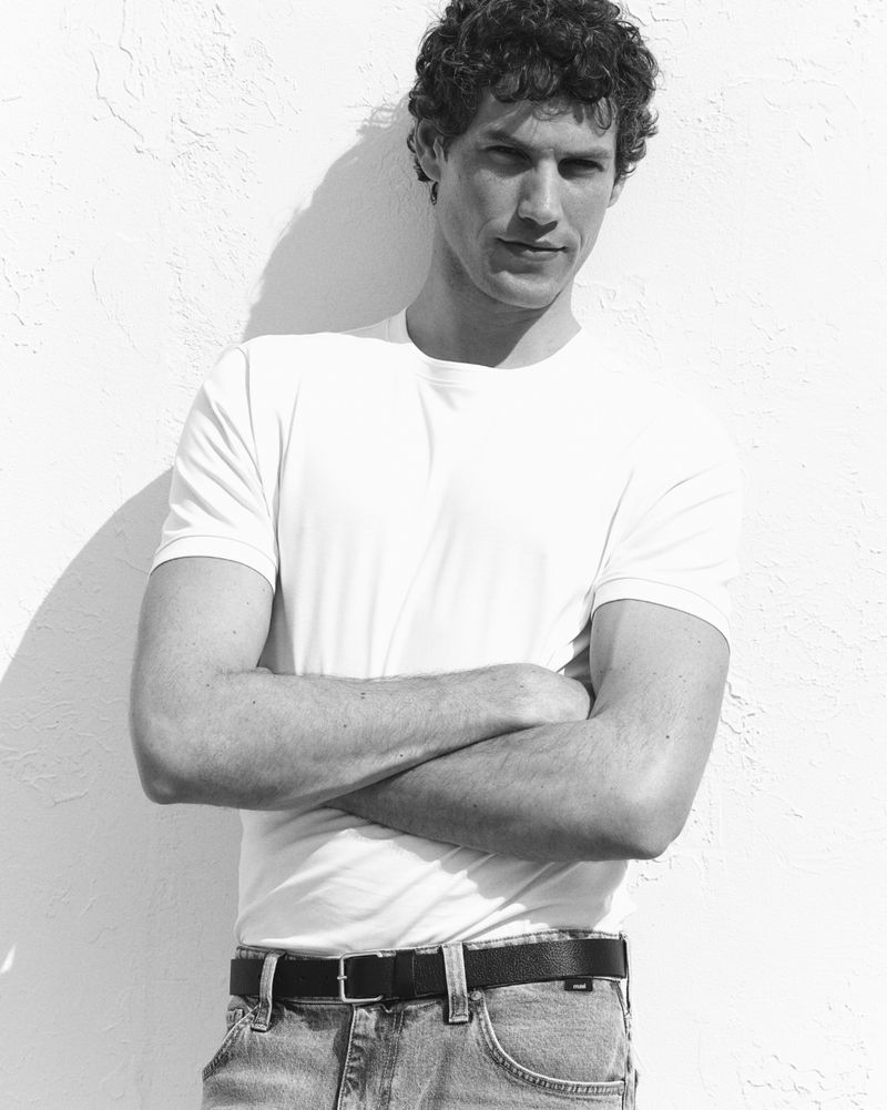 Paul Hupfer posing in a white crewneck t-shirt and denim jeans against a textured wall in black and white.