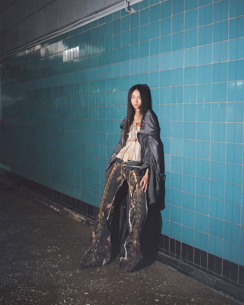 Model standing against a dark turquoise tiled wall wearing an oversized grey jacket and patterned trousers
