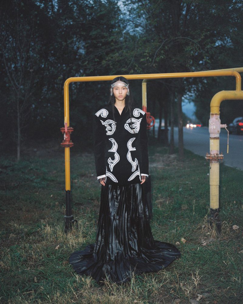 Model wearing an embroidered black coat and pleated black maxi skirt standing outdoors in front of yellow utility pipes at dusk.