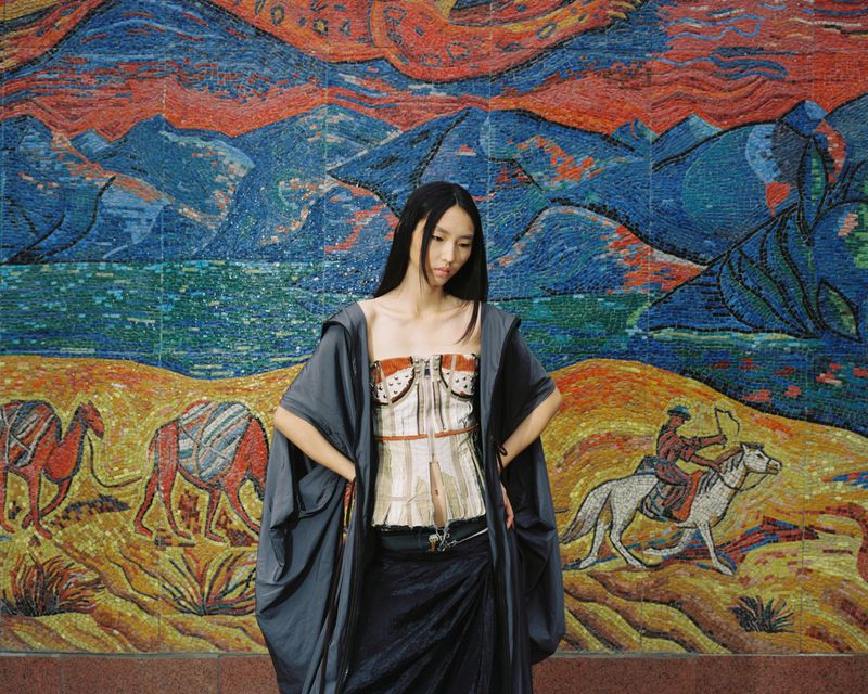 Model standing against a large mosaic mural depicting mountains and riders in red and blue tones