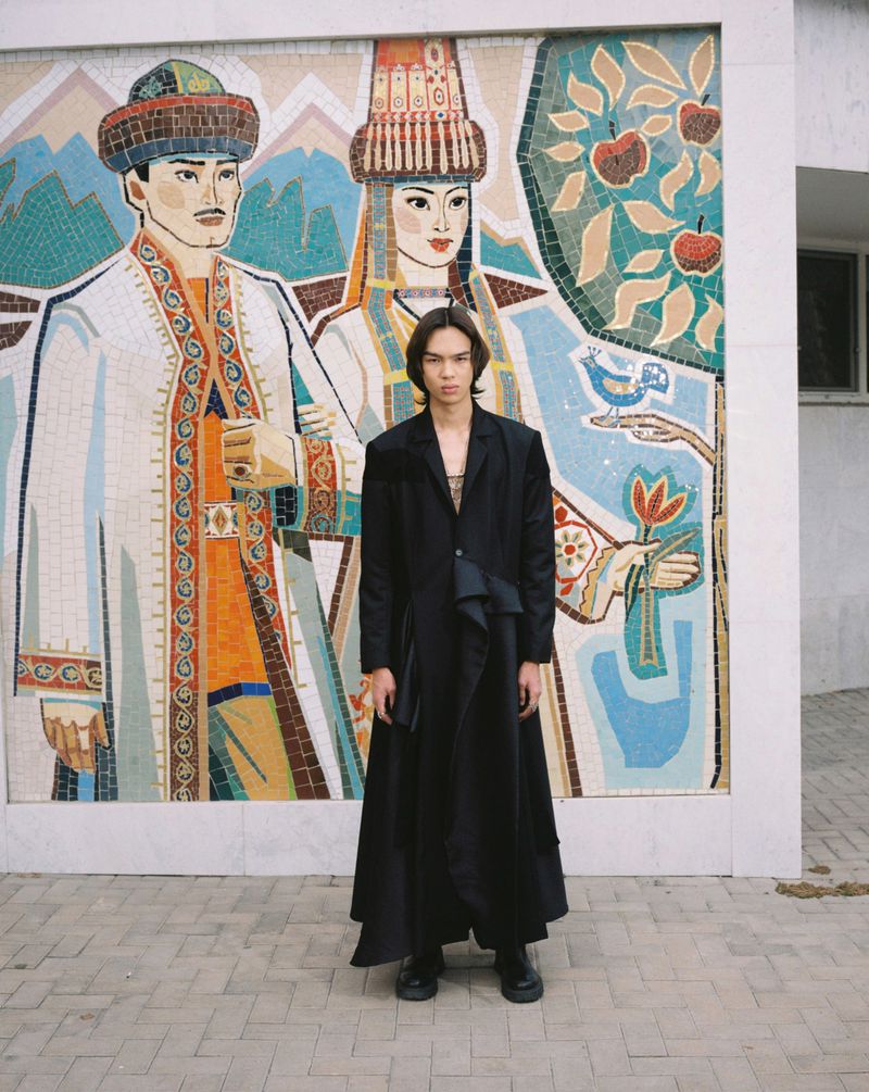 Model Aldiyar Amangeldy stands in a long black coat before a colorful tiled Central Asian mosaic mural.