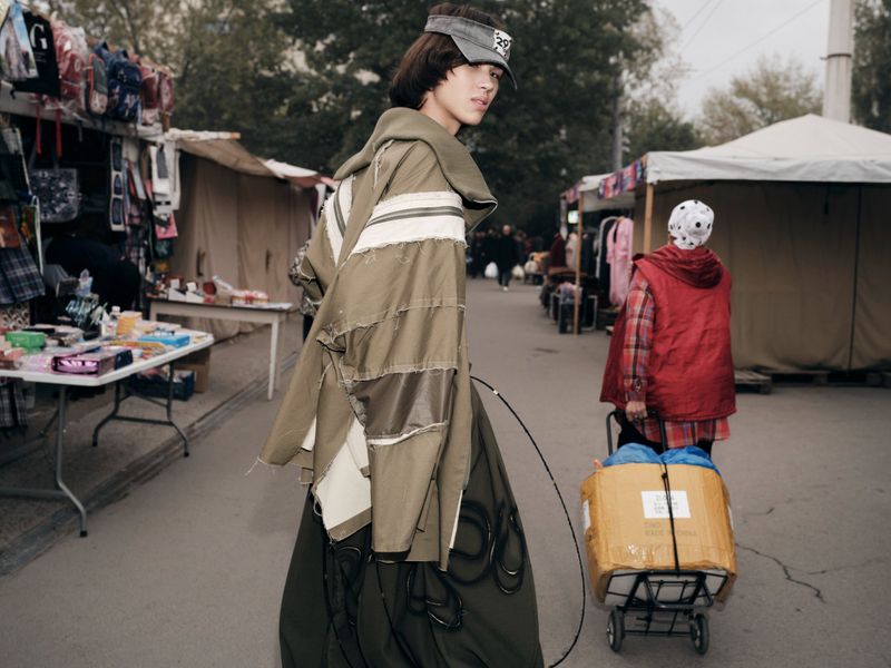 Model Aldiyar Amangeldy looks back in a deconstructed olive green jacket and visor at an outdoor market.
