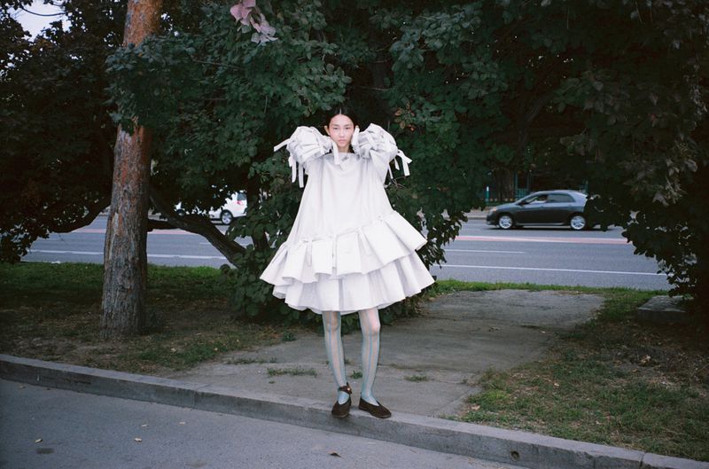 Model wearing a voluminous tiered white dress and Mary Jane flats standing on a sidewalk near lush green trees.
