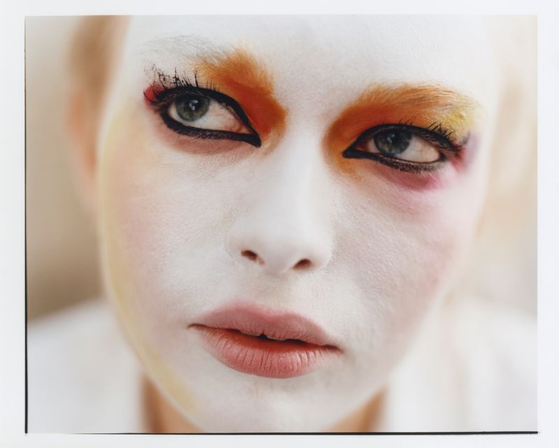 Close-up beauty portrait of a model with white face paint, graphic black eyeliner, and vibrant orange eye makeup.