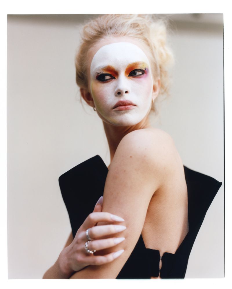 Model Yana Palkhouskaya with white mask-like face paint and orange eye shadow wearing a sculptural black top in an editorial portrait.