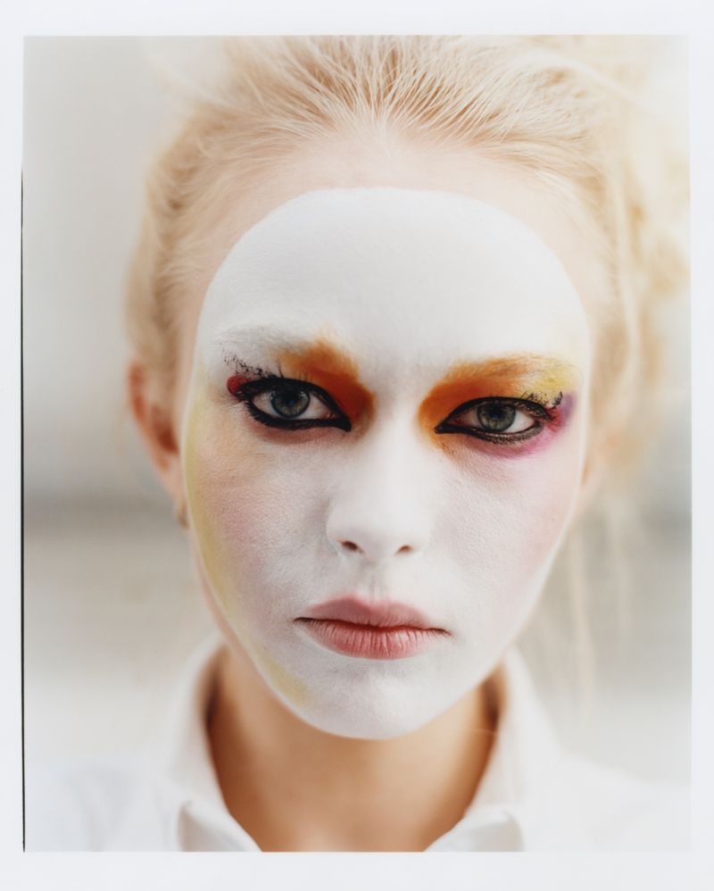 Close-up beauty portrait of model Yana Palkhouskaya wearing theatrical white face paint and colorful eye makeup with a white shirt.