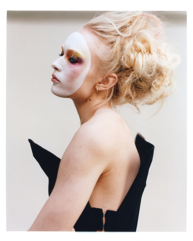 Model with avant-garde white face makeup and blonde updo wearing a structured black top