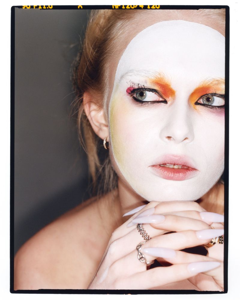 Editorial beauty close-up of model Yana Palkhouskaya with white face paint, colorful eye makeup, and long pointed nails.