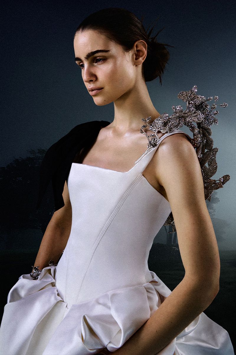 Woman in white formal dress with ornamental shoulder piece poses in a misty outdoor setting, elegant fashion photography.