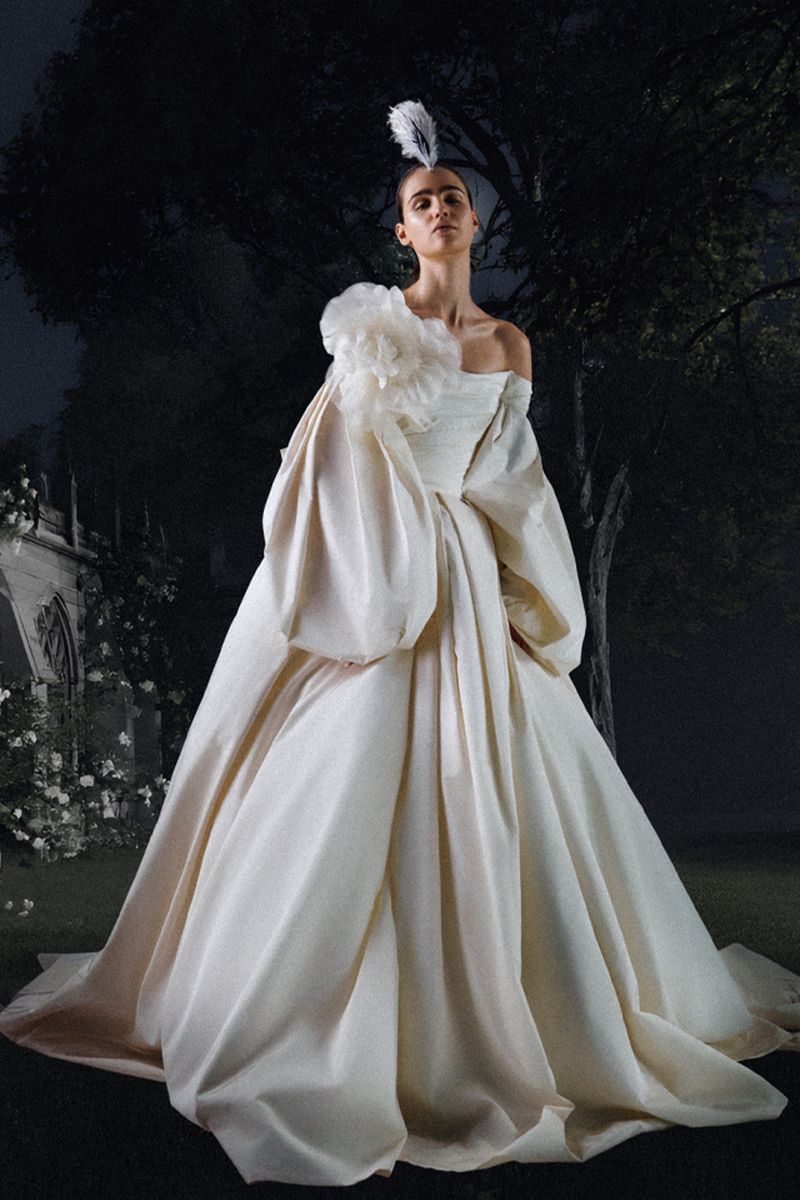 An adult female model poses in a formal off-the-shoulder white gown with large puffy sleeves and a white feather headpiece in a dark outdoor scene.