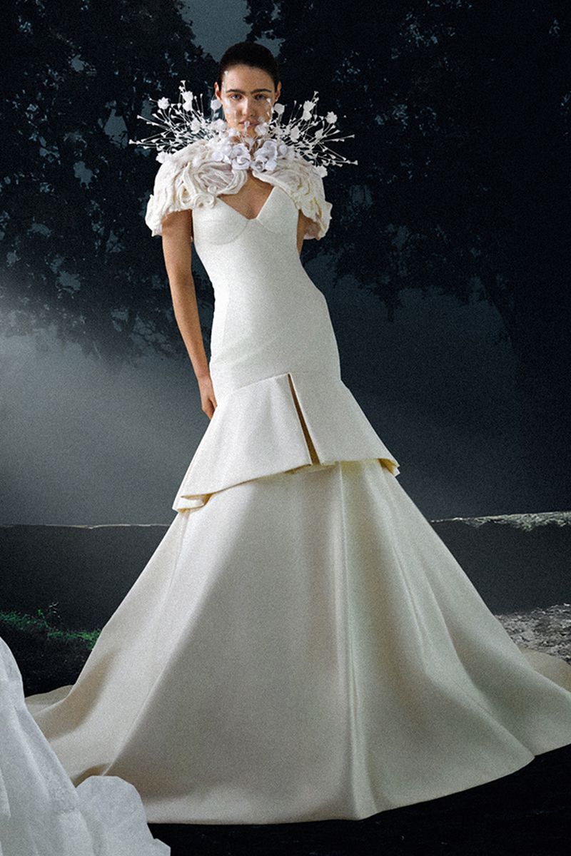 A woman in a white gown poses in front of a moody, dark background. The gown is floor-length with tiers, and she wears a flower collar.