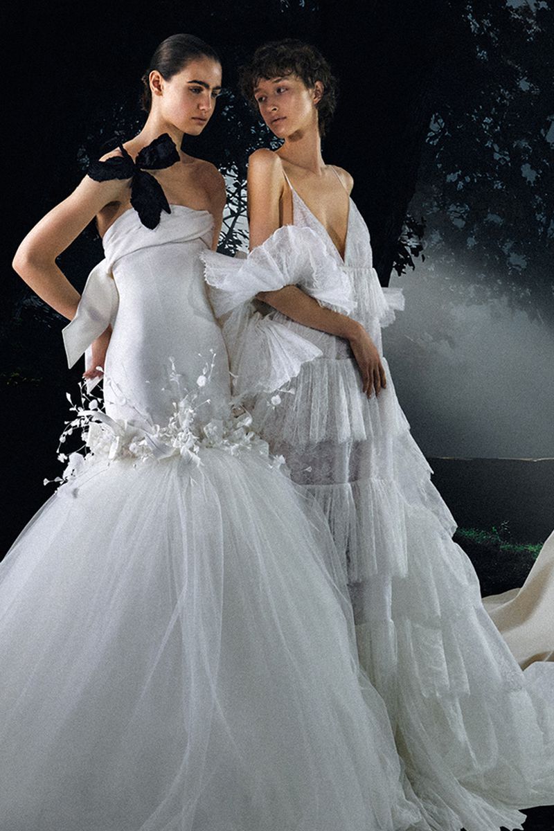 Two women in elegant white gowns with decorative details pose against a dark background with tree silhouettes in a fashion shot.