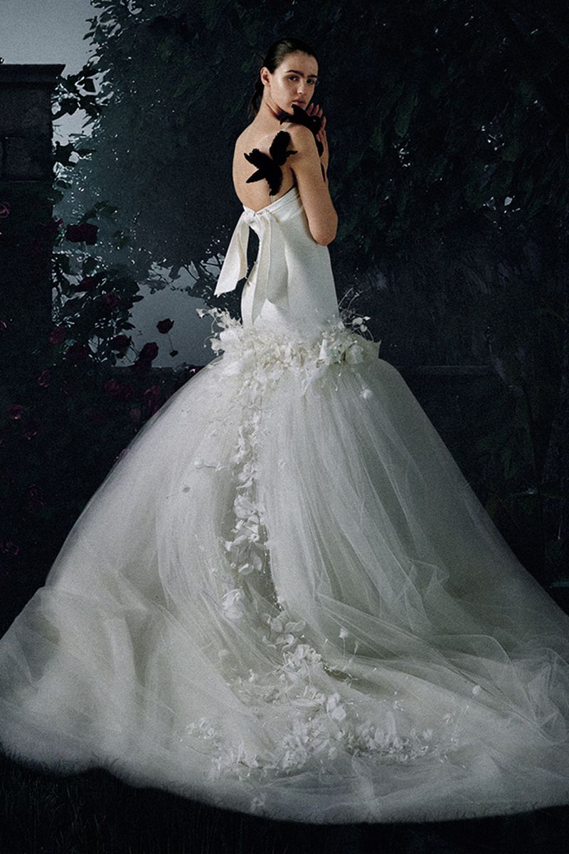 Woman in a strapless white wedding gown with a long train adorned with white floral accents, standing against a dark floral background.
