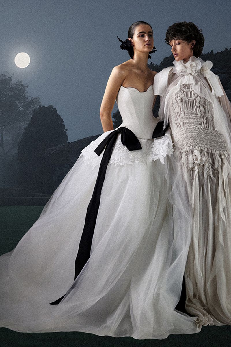 Two fashion models in white gowns pose outdoors under a full moon in an artistic setting, one gown with a black bow.