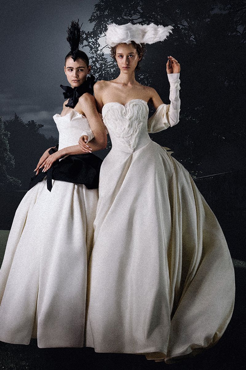 Two women in white gowns pose in a fashion shot with a dark, surreal background featuring trees and dramatic lighting.