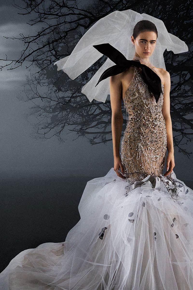 Woman in beaded sleeveless gown with white tulle skirt, and large black bow detail, poses against a dark grey forest background.