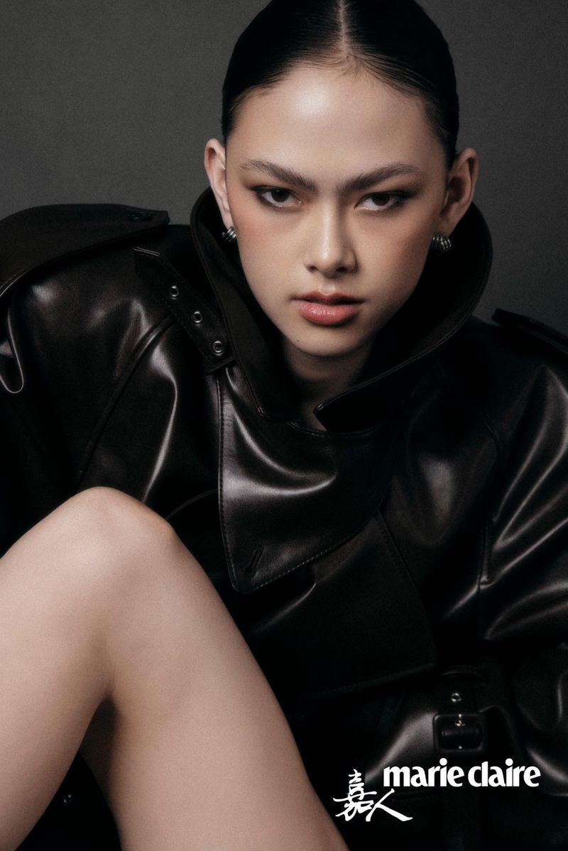 East Asian female model in an oversized black leather coat posing against a dark grey seamless studio backdrop.