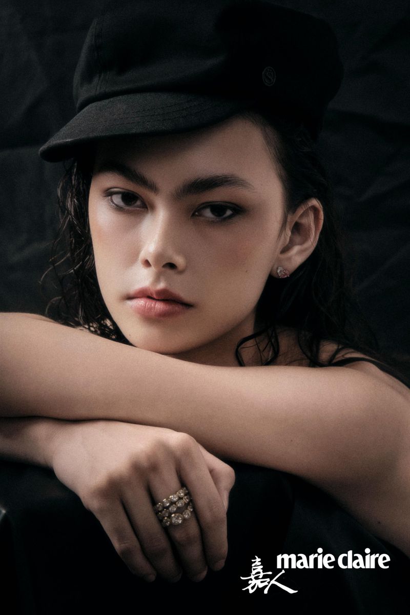 Model wearing a black cap and layered diamond ring resting her chin on crossed arms against a dark backdrop.