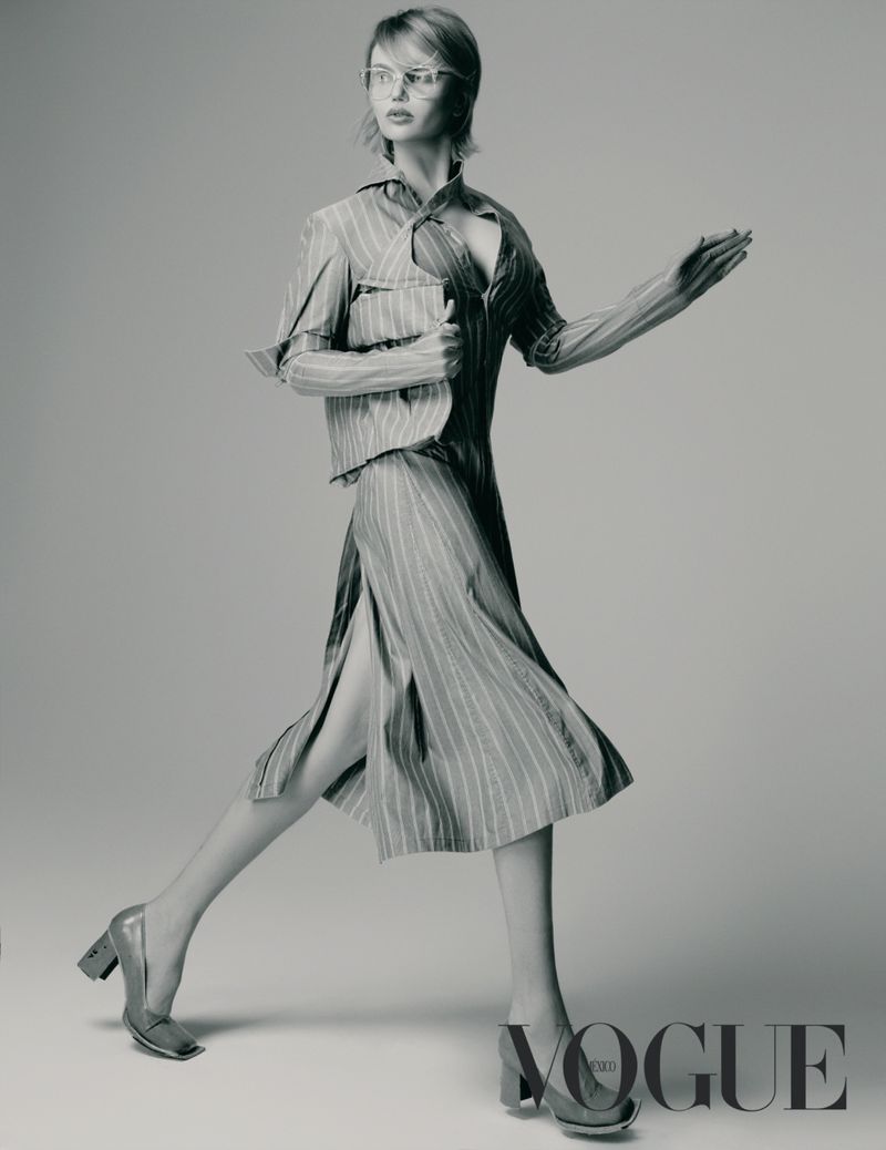 Elena Matei walks dynamically across a studio wearing a striped deconstructed dress and eyeglasses in a monochrome editorial portrait.