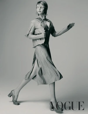 Elena Matei walks dynamically across a studio wearing a striped deconstructed dress and eyeglasses in a monochrome editorial portrait.