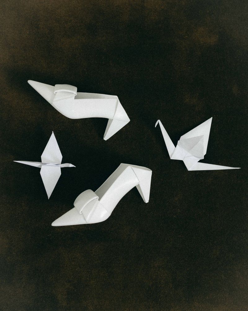 White pointed-toe sculptural high heel shoes arranged with folded origami paper cranes on a dark textured background.