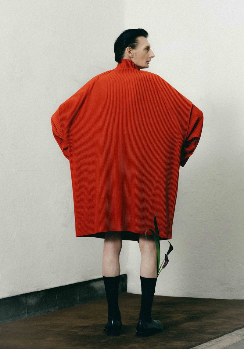 Man seen from behind wearing an oversized ribbed red knit tunic with black socks and shoes against a studio corner wall.
