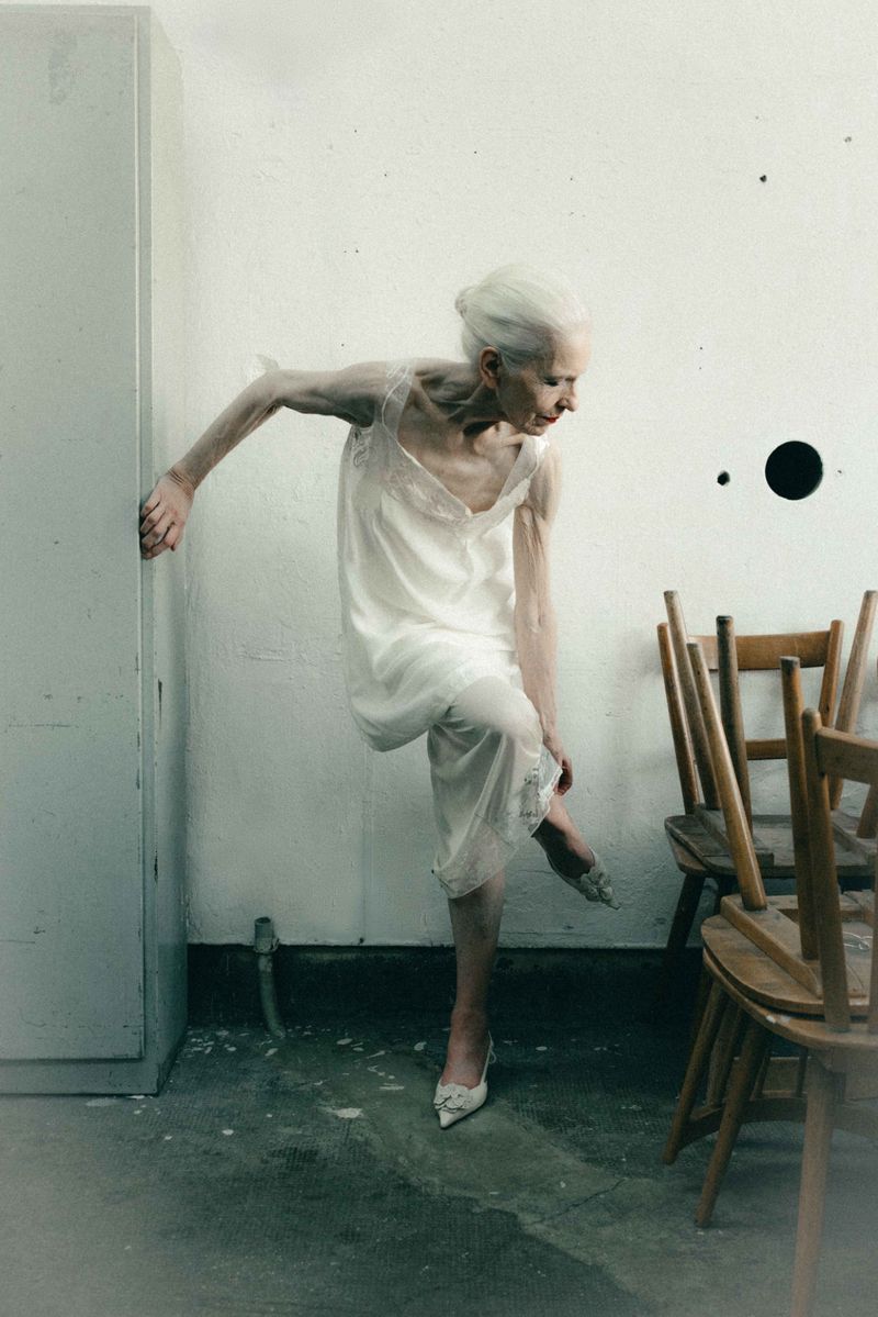 Senior woman with white hair wearing an off-white slip dress and pointed heels in a sparse room beside wooden chairs.