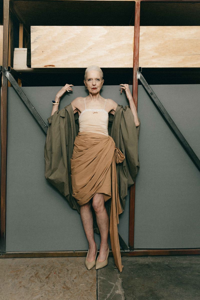 Elderly woman with slicked-back white hair posing in a draped tan skirt and oversized khaki jacket against grey studio set flats.