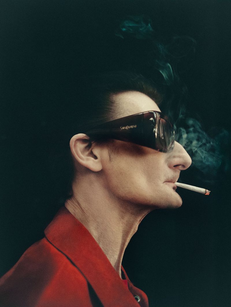 Profile portrait of a man wearing oversized dark sunglasses and a red jacket while smoking a lit cigarette against a dark backdrop.