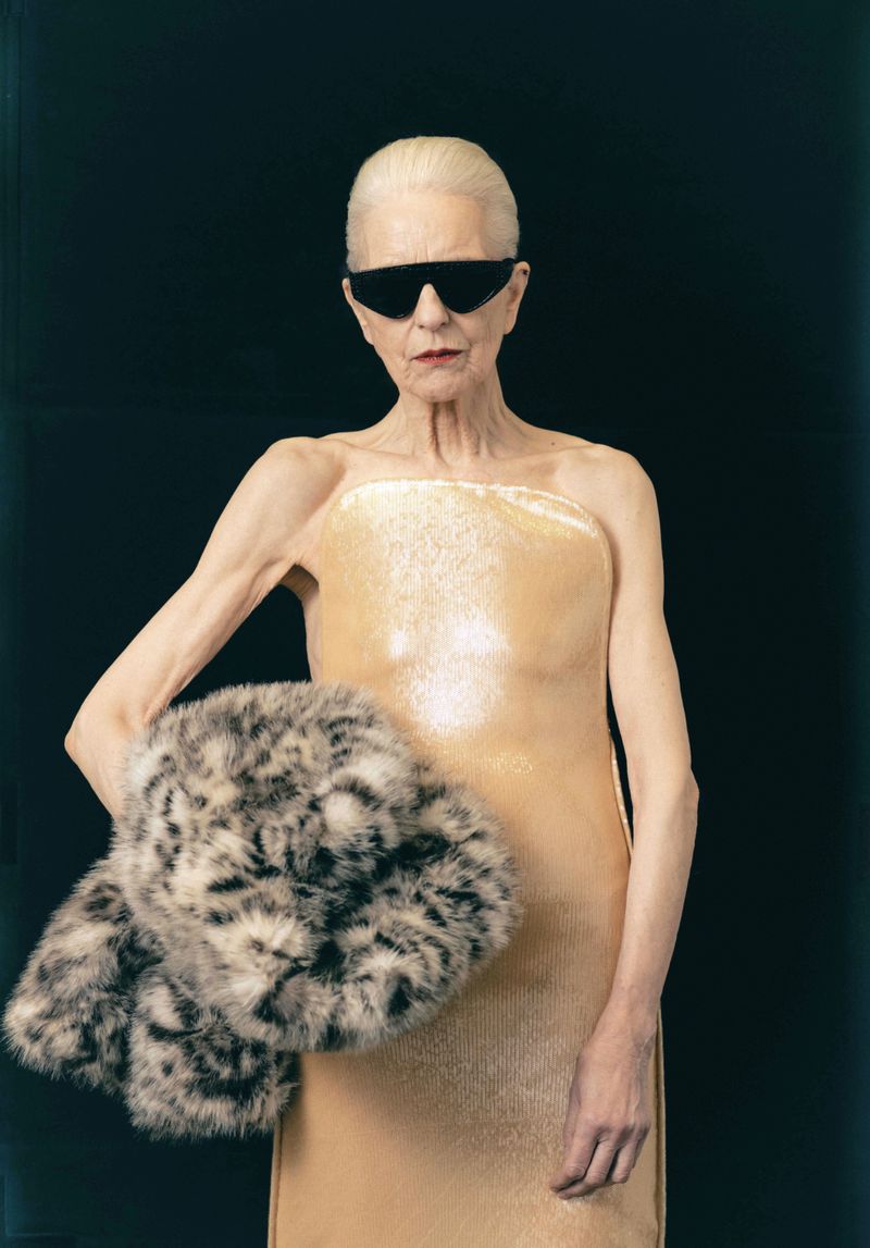 Elderly woman wearing black angular sunglasses and a strapless beige gown holding a spotted fur stole against a black backdrop.