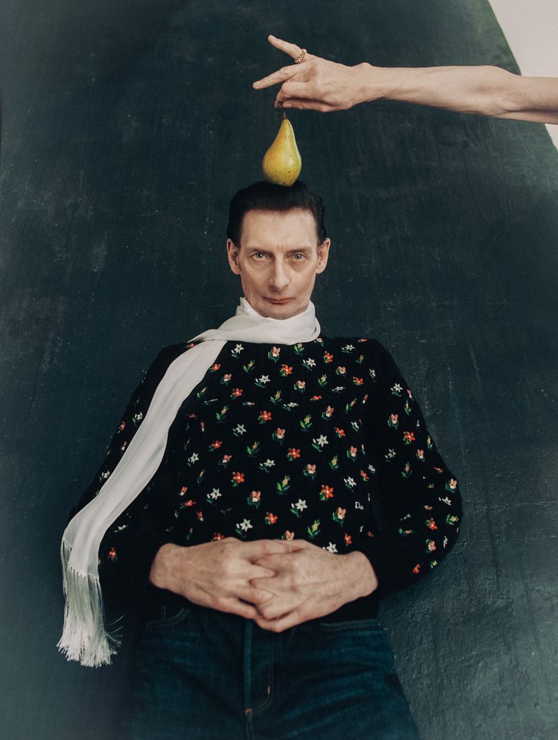 Model Steve Morell lying down in a floral knit sweater and white fringed scarf with a yellow pear held above his head.
