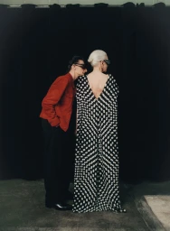 Model in a black and white polka dot gown standing with a man wearing a red jacket against a black curtain backdrop.