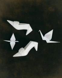 White pointed-toe sculptural high heel shoes arranged with folded origami paper cranes on a dark textured background.