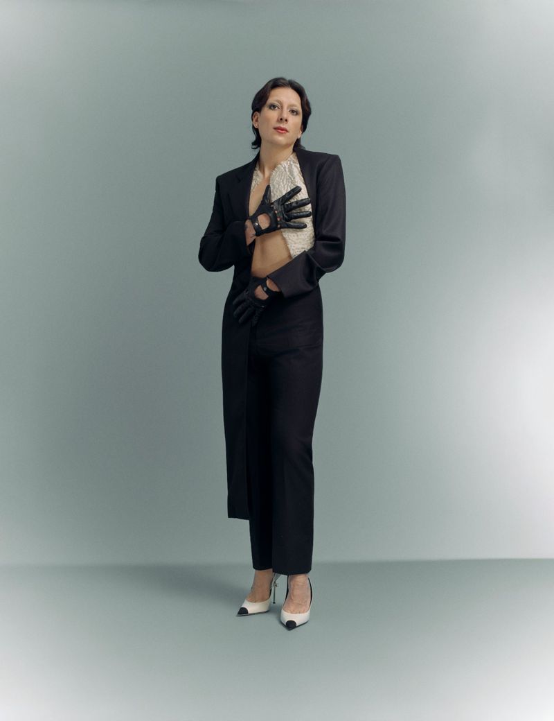 Mason Alexander Park posing in a black tailored suit jacket, trousers, cutout gloves, and two-tone heels against a grey studio backdrop.