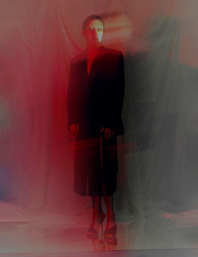 Mason Alexander Park posing in a tailored black blazer and skirt under red atmospheric lighting.