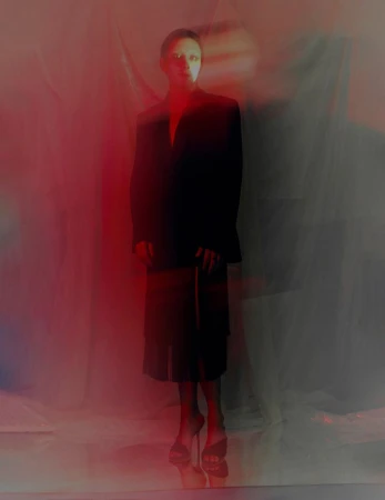 Mason Alexander Park posing in a tailored black blazer and skirt under red atmospheric lighting.