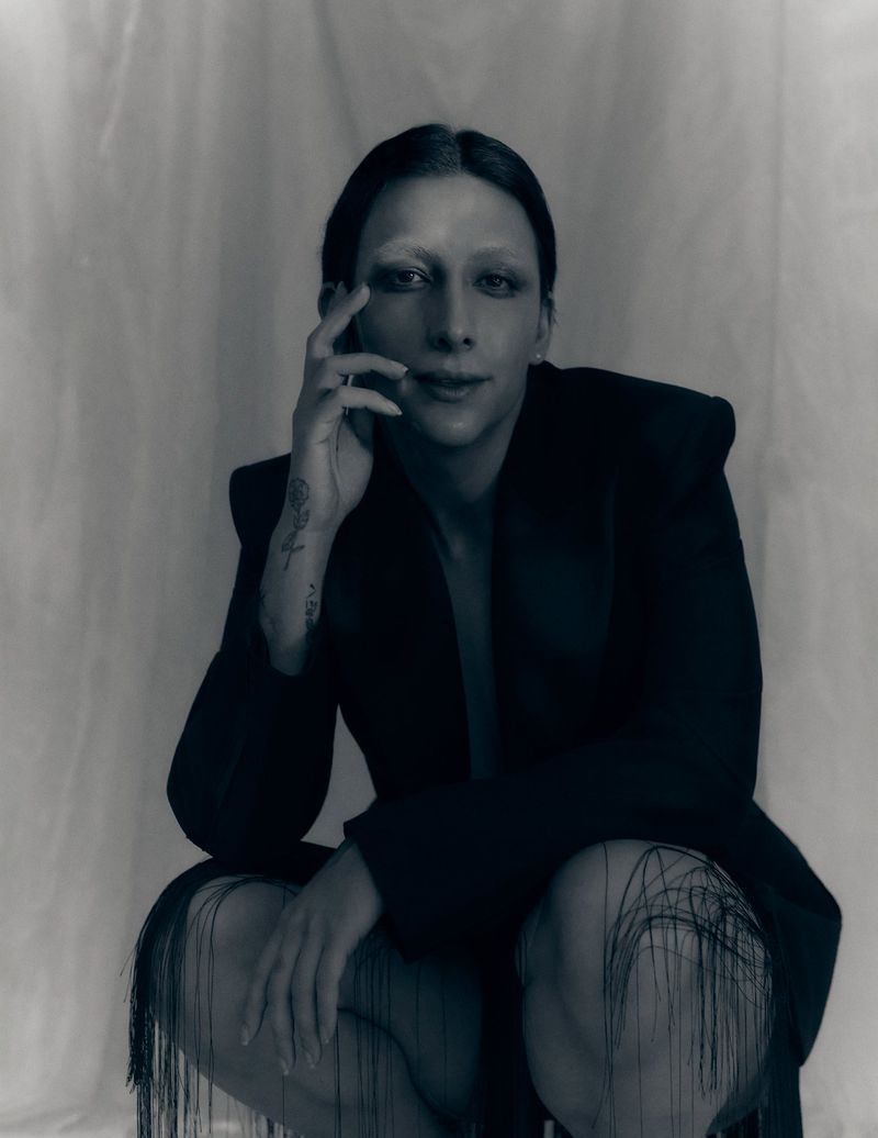 Mason Alexander Park crouching in a black tailored blazer with fringe accents against a softly draped studio backdrop.