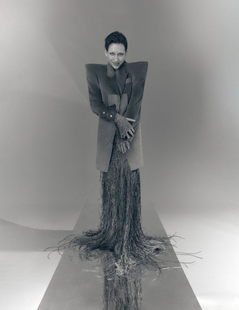 Mason Alexander Park posing in an avant-garde exaggerated shoulder coat and trailing fringe skirt on a reflective studio runway.