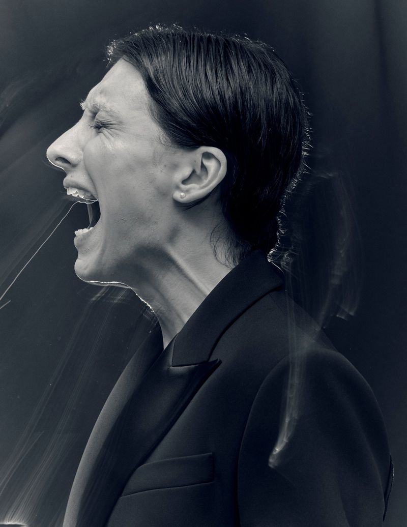 Monochrome profile portrait of Mason Alexander Park shouting with mouth open, wearing a black tailored blazer against a dark background.