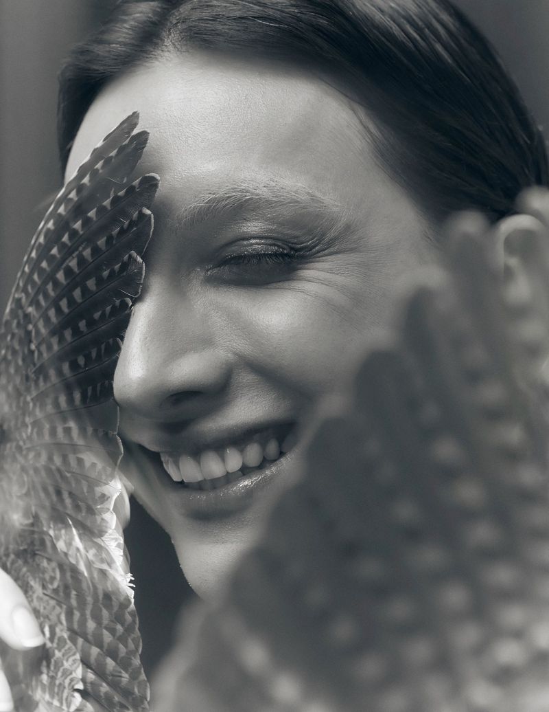 Black and white close-up portrait of Mason Alexander Park smiling behind a patterned feathered wing.