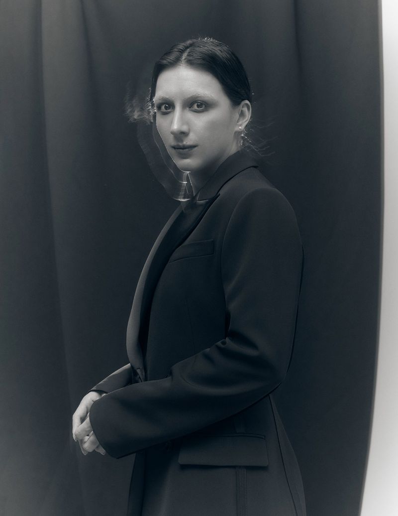 Monochrome portrait of Mason Alexander Park wearing a black tailored blazer against a dark draped fabric studio backdrop.