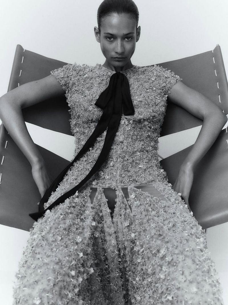 Model Mar G seated in a leather sling chair wearing a textured appliqué dress with a black neck ribbon against a white background.