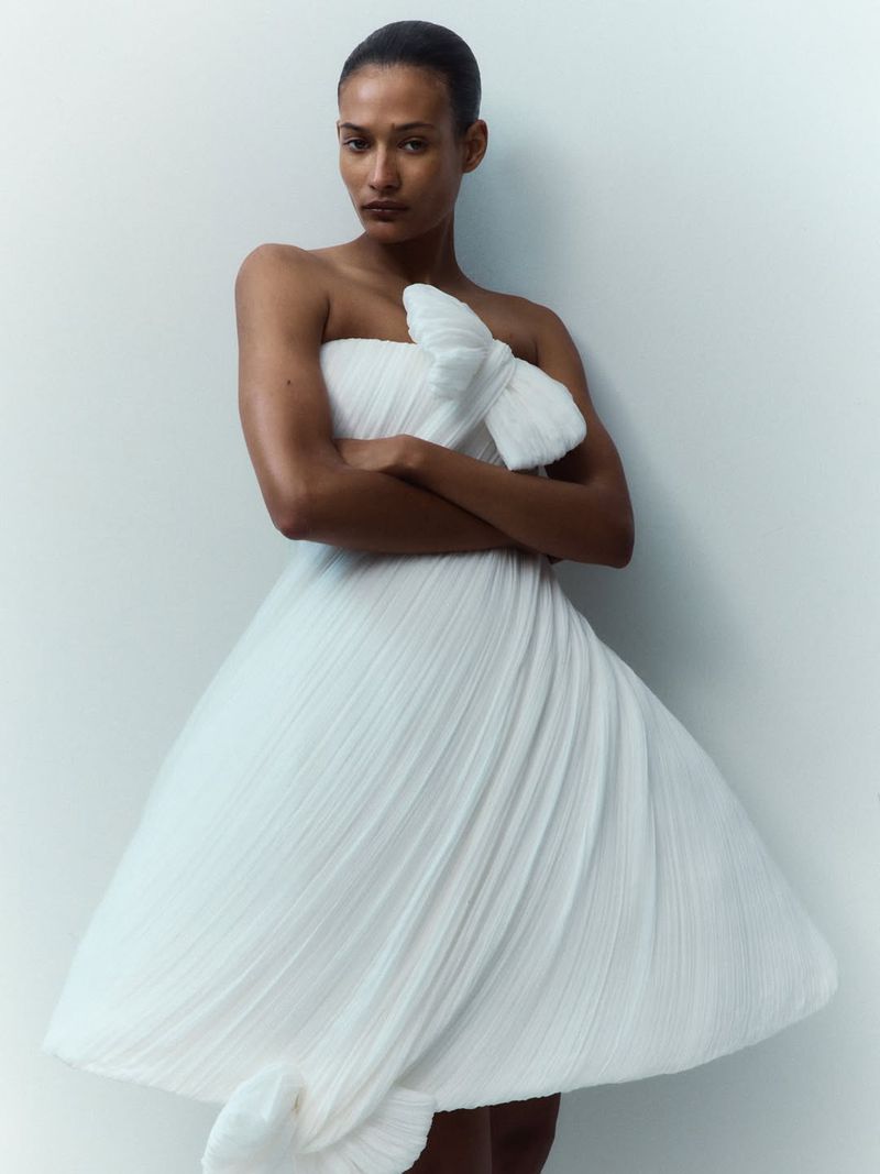Model Mar G wearing a sculptural strapless white pleated dress with arms crossed against a neutral studio backdrop.